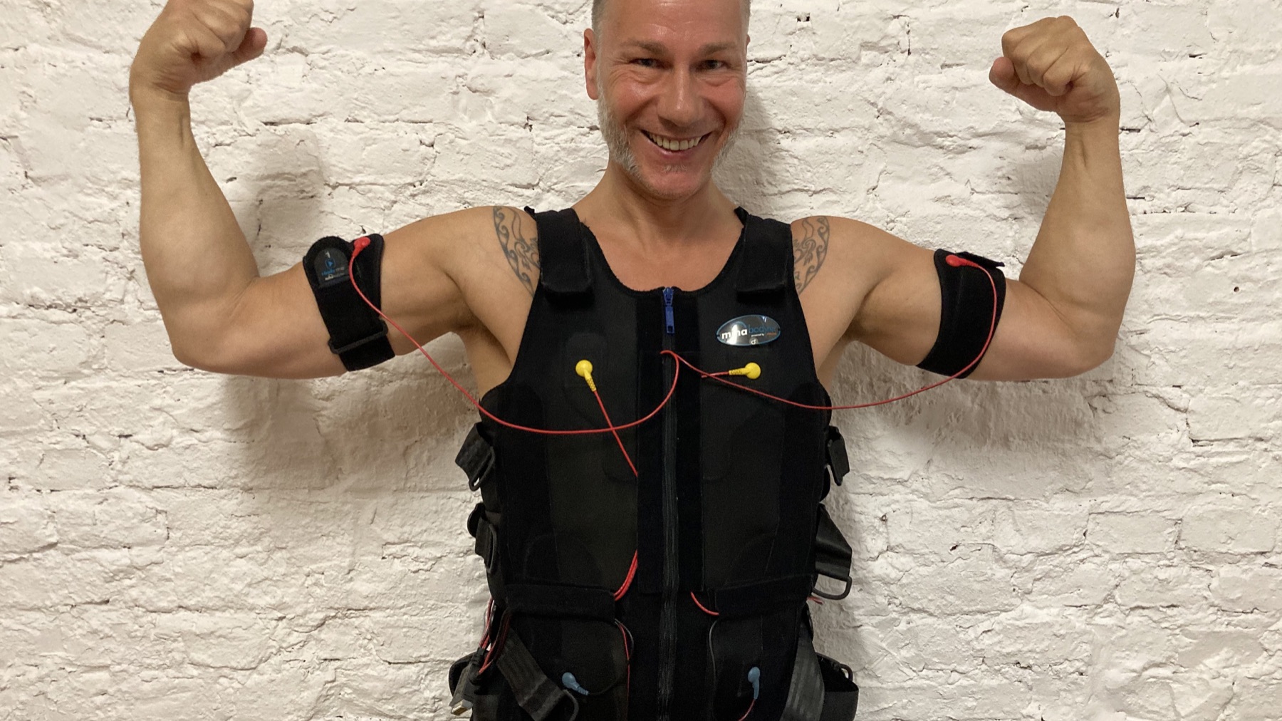 Peter lächelt im EMS-Trainingsanzug vor einer hellen Backsteinwand